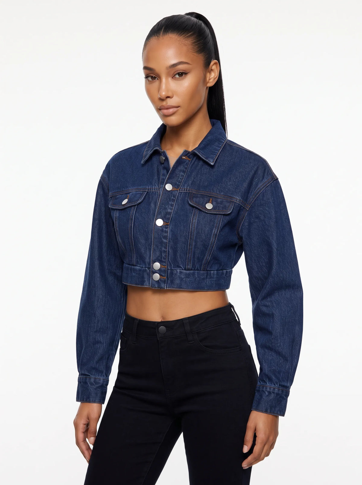 This is a deep indigo blue denim jacket features an ultra-cropped silhouette with long sleeves, tailored shoulder seams, and classic buttoned cuffs. It includes a traditional point collar and a full front placket detailed with smooth, metallic silver shank buttons.
