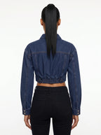 This is a deep indigo blue denim jacket features an ultra-cropped silhouette with long sleeves, tailored shoulder seams, and classic buttoned cuffs. It includes a traditional point collar and a full front placket detailed with smooth, metallic silver shank buttons.