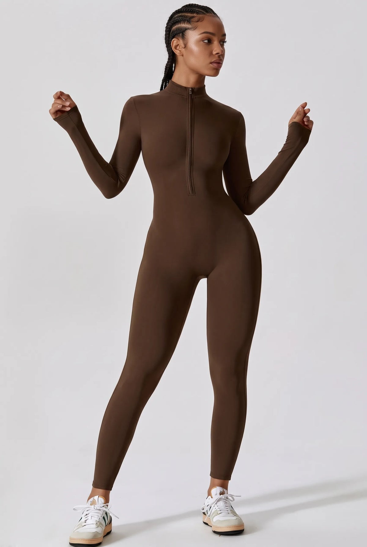 A model wearing a brown long-sleeve mock-neck full-zip bodysuit, paired with white sneakers, standing against a light gray studio background.