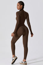 A woman modeling a tight-fitting, long-sleeve, mock-neck, high-waisted brown bodysuit from the back, paired with white and black sneakers, against a light gray studio background.