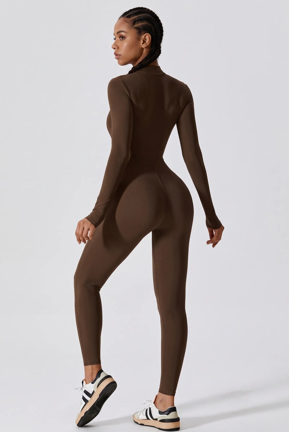 A woman modeling a tight-fitting, long-sleeve, mock-neck, high-waisted brown bodysuit from the back, paired with white and black sneakers, against a light gray studio background.
