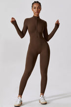 A model wearing a brown long-sleeve mock-neck full-zip bodysuit, paired with white sneakers, standing against a light gray studio background.