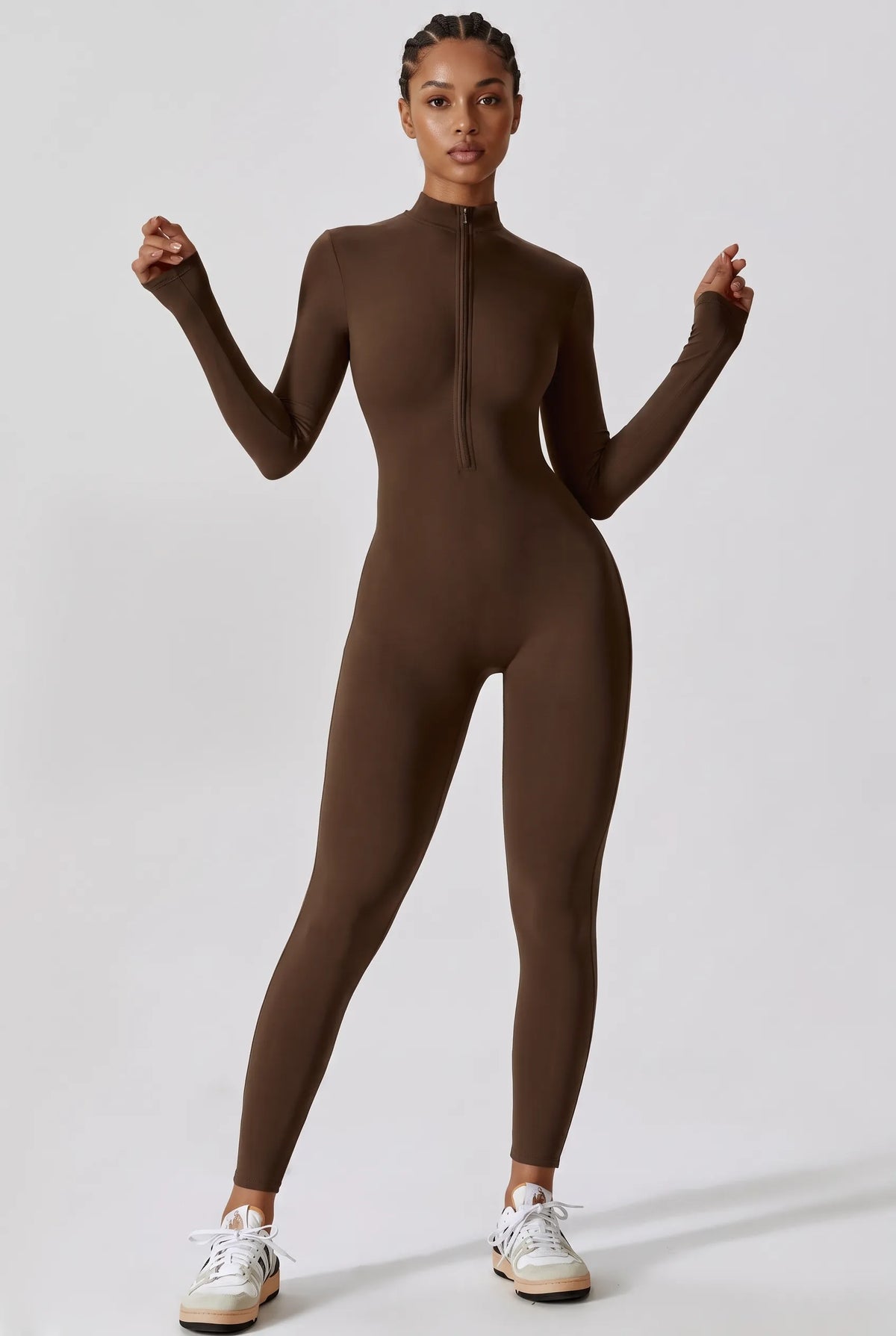A model wearing a brown long-sleeve mock-neck full-zip bodysuit, paired with white sneakers, standing against a light gray studio background.
