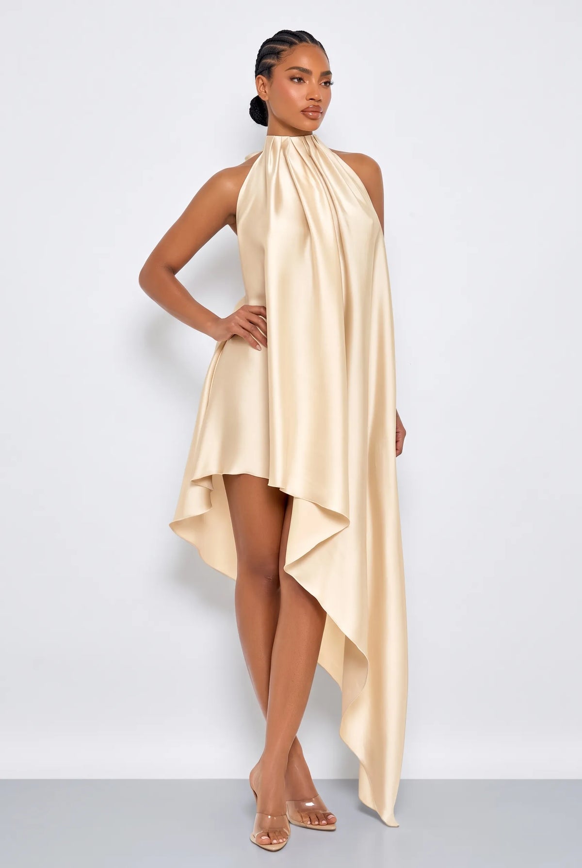 A model wearing a champagne gold high-neck halter dress with a draped, asymmetrical flowy skirt, posed against a light gray studio background.