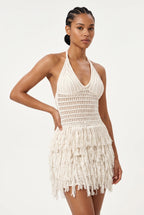 A woman modeling a cream-colored crochet halter mini dress with a fringed hem against a plain white studio background.
