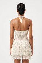 A model shown from the back wearing a cream-colored crochet halter mini dress with a tiered fringe skirt, set against a plain white studio background.