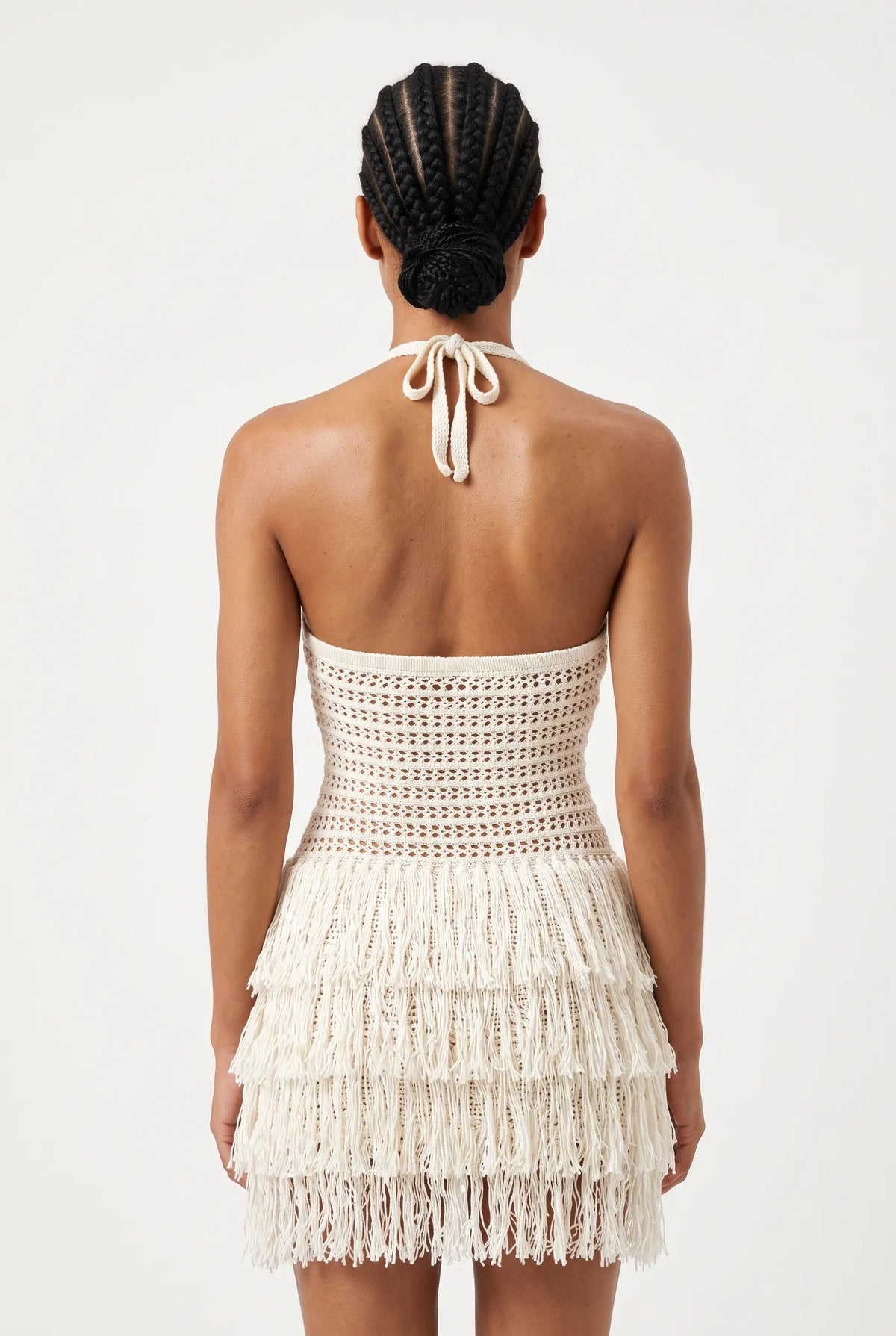 A model shown from the back wearing a cream-colored crochet halter mini dress with a tiered fringe skirt, set against a plain white studio background.