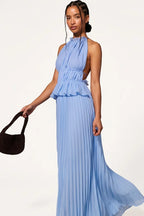 A woman models a light blue halter-neck maxi dress with a ruffled peplum waist and pleated skirt, holding a dark brown woven handbag against a plain white studio background.