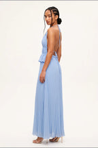 A woman modeling a light blue pleated maxi dress with a halter neck, open back, and peplum waist detail, shown from a side-back angle against a plain white studio background.