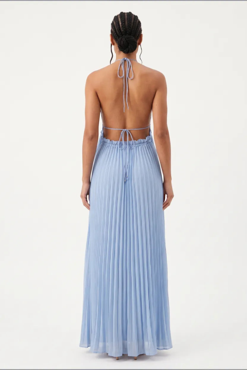 A woman viewed from the back wearing a light blue pleated maxi dress with an open back and tie detail at the neck, photographed against a plain white studio background.