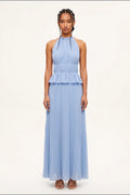 A model wearing a light blue sleeveless halter-neck maxi dress with a ruffled waist and pleated skirt, standing against a plain off-white studio background.