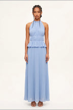 A model wearing a light blue sleeveless halter-neck maxi dress with a ruffled waist and pleated skirt, standing against a plain off-white studio background.