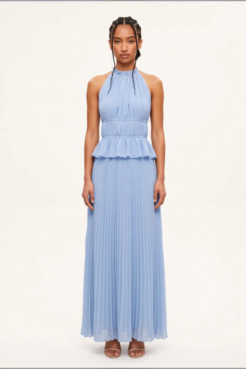 A model wearing a light blue sleeveless halter-neck maxi dress with a ruffled waist and pleated skirt, standing against a plain off-white studio background.