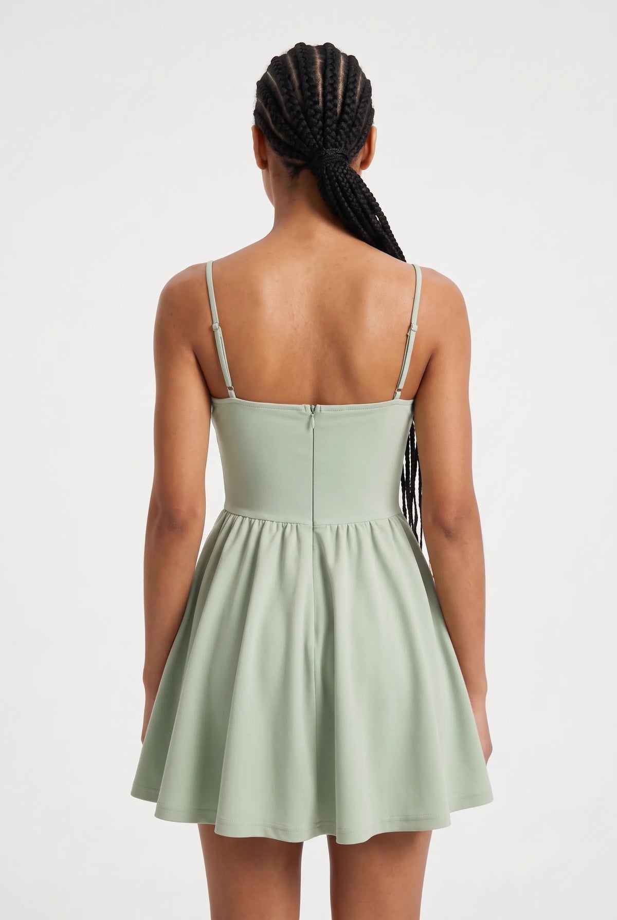 A woman modeling a sage green spaghetti-strap mini dress with a ruched bodice and flared skirt against a plain light background.