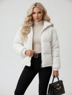 Woman wearing a white puffer jacket, beige turtleneck, black pants, and holding a black handbag on a light gray background.