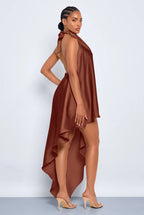 A woman modeling a brown satin halter-neck high-low dress with an open back, shown from a side-back angle against a light gray studio background.