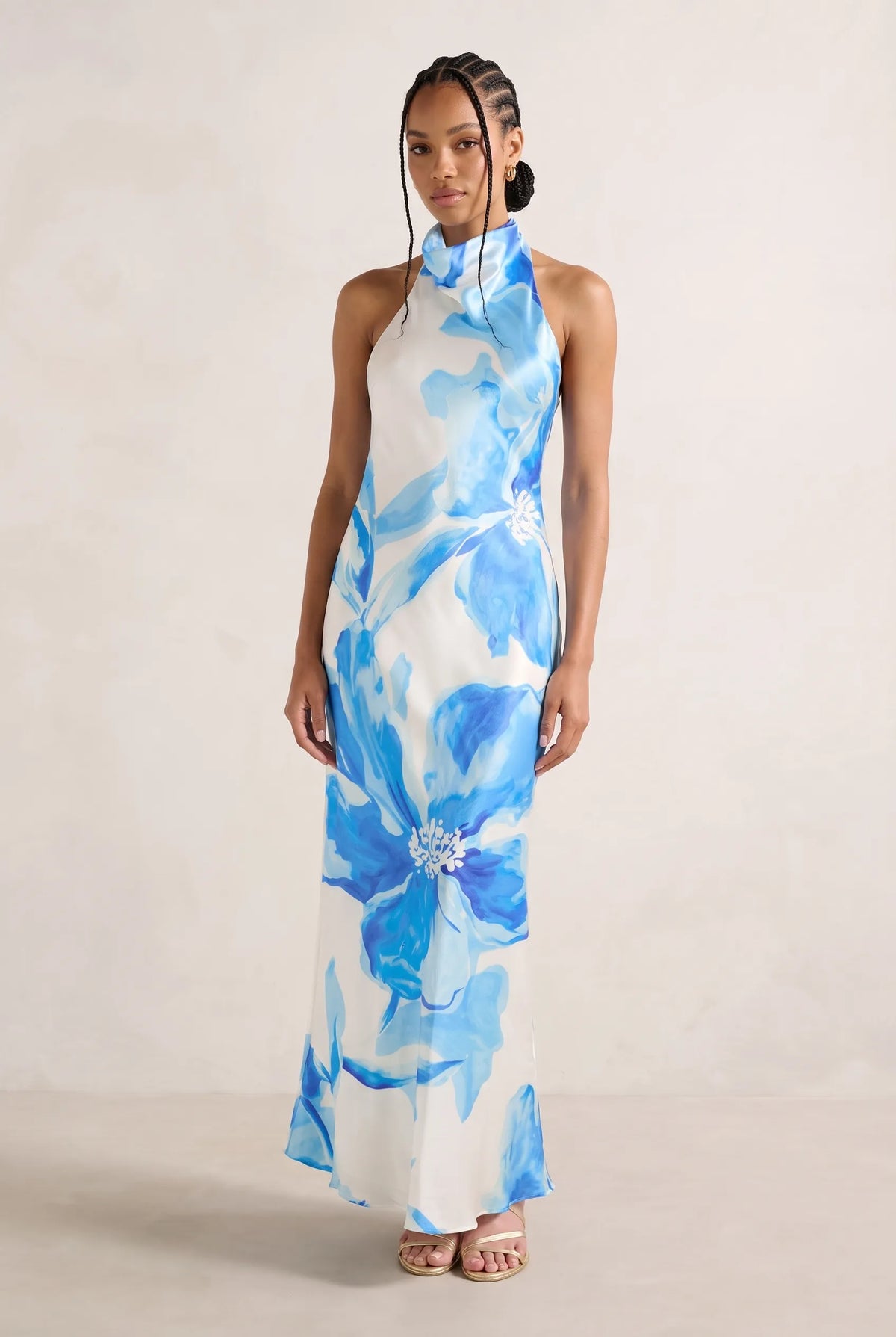 A woman modeling a sleeveless high-neck maxi dress with a white base and large blue watercolor-style floral print, standing against a plain light beige studio backdrop.