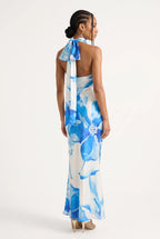 A woman modeling a white halter maxi dress with a blue watercolor floral print, shown from the back against a plain white studio background.