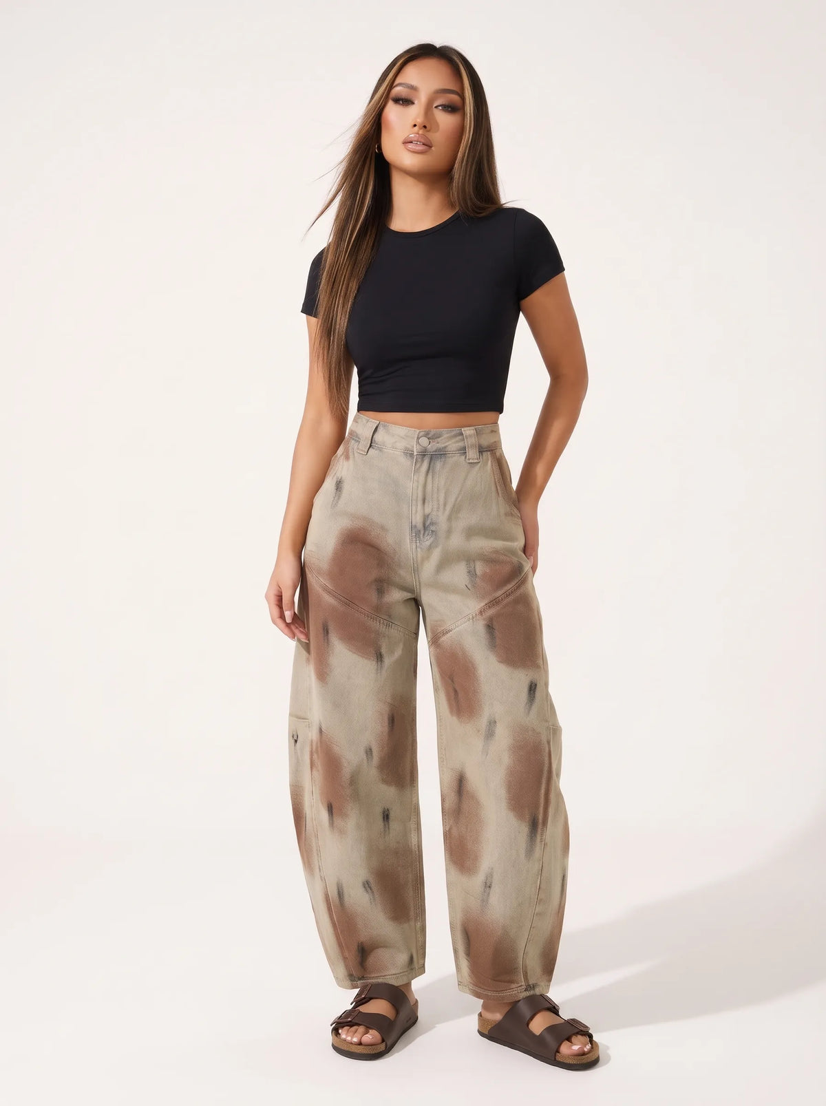 A model wearing a pair of high-waisted, curved barrel-leg jeans is crafted from a vintage-washed light khaki or beige denim adorned with an abstract, artistic spray-paint effect featuring mottled splotches of warm rust brown and distressed charcoal black.