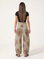 A model wearing a pair of high-waisted, curved barrel-leg jeans is crafted from a vintage-washed light khaki or beige denim adorned with an abstract, artistic spray-paint effect featuring mottled splotches of warm rust brown and distressed charcoal black.