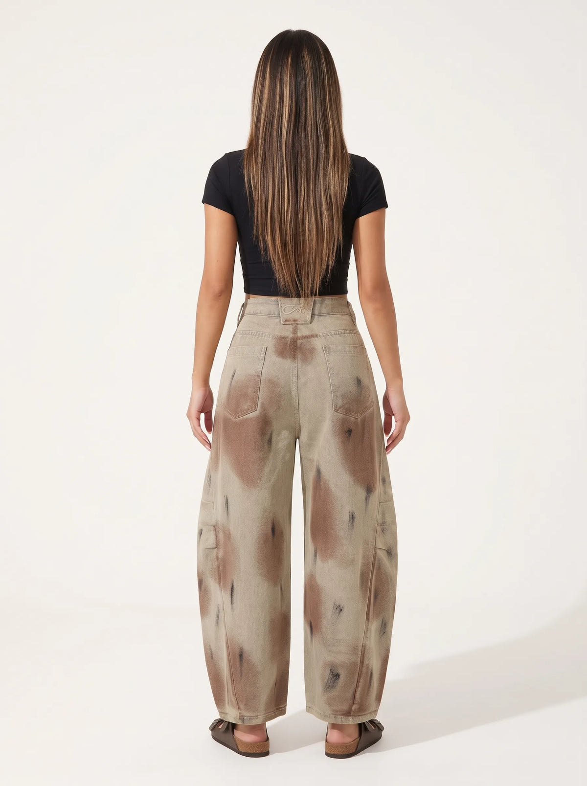 A model wearing a pair of high-waisted, curved barrel-leg jeans is crafted from a vintage-washed light khaki or beige denim adorned with an abstract, artistic spray-paint effect featuring mottled splotches of warm rust brown and distressed charcoal black.