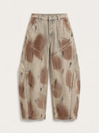 A  pair of high-waisted, curved barrel-leg jeans is crafted from a vintage-washed light khaki or beige denim adorned with an abstract, artistic spray-paint effect featuring mottled splotches of warm rust brown and distressed charcoal black.