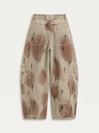 A  pair of high-waisted, curved barrel-leg jeans is crafted from a vintage-washed light khaki or beige denim adorned with an abstract, artistic spray-paint effect featuring mottled splotches of warm rust brown and distressed charcoal black.