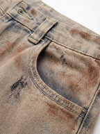 A  pair of high-waisted, curved barrel-leg jeans is crafted from a vintage-washed light khaki or beige denim adorned with an abstract, artistic spray-paint effect featuring mottled splotches of warm rust brown and distressed charcoal black.