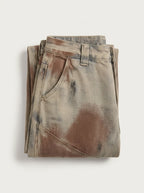 A  pair of high-waisted, curved barrel-leg jeans is crafted from a vintage-washed light khaki or beige denim adorned with an abstract, artistic spray-paint effect featuring mottled splotches of warm rust brown and distressed charcoal black.