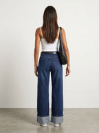 a model wearing a simple White thin-strap cropped camisole top paired with high-waisted dark blue wash wide-leg jeans featuring prominent wide turned-up cuffed hems. Styled with sleek black sunglasses, a structured black leather shoulder bag, and White  snicker
