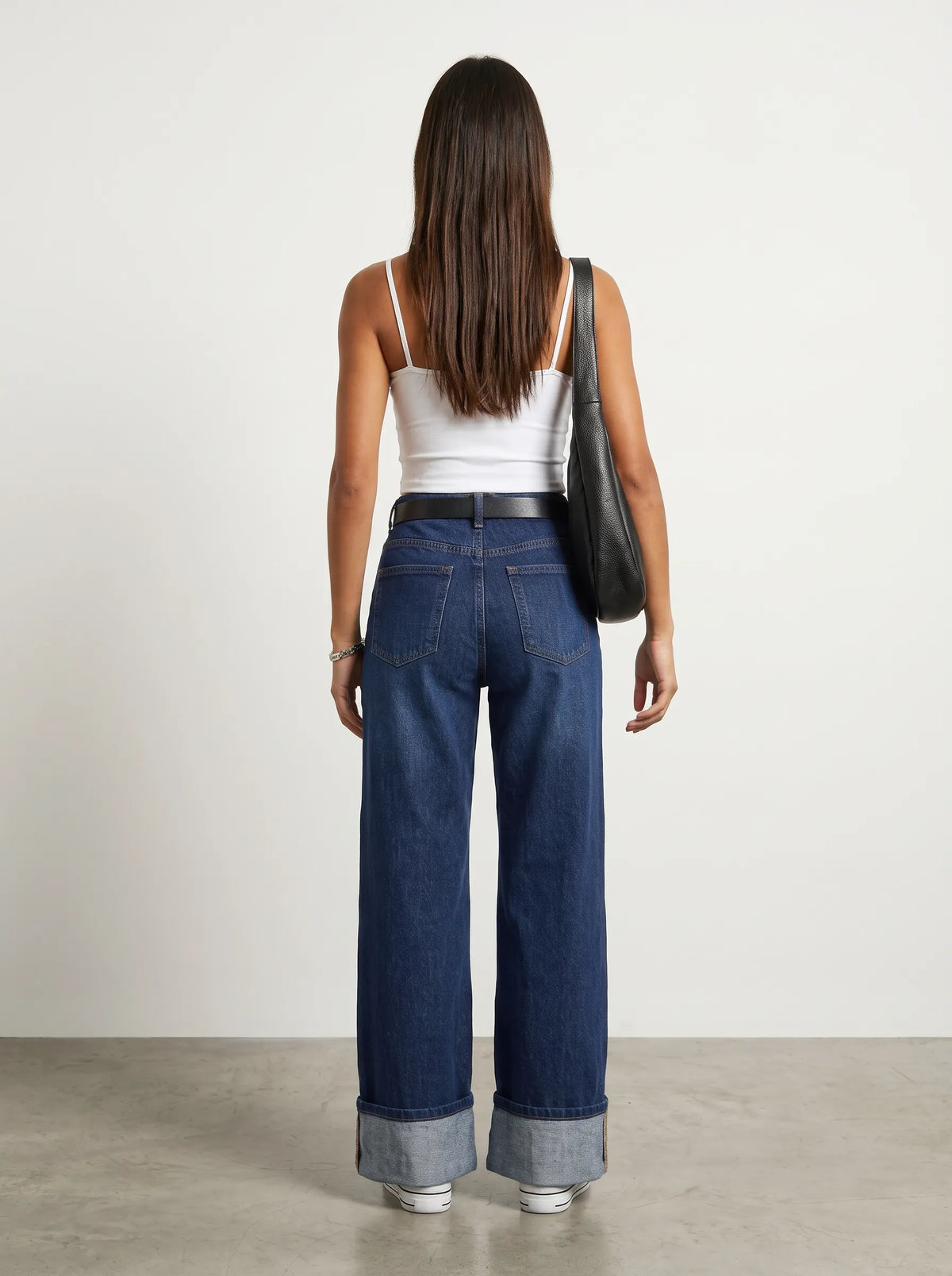 a model wearing a simple White thin-strap cropped camisole top paired with high-waisted dark blue wash wide-leg jeans featuring prominent wide turned-up cuffed hems. Styled with sleek black sunglasses, a structured black leather shoulder bag, and White  snicker