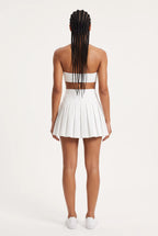 A model viewed from the back wearing a white two-piece outfit consisting of a strapless bandeau-style crop top with a horizontal back cutout and a matching high-waisted pleated mini skirt, paired with white sneakers against a plain off-white studio background.