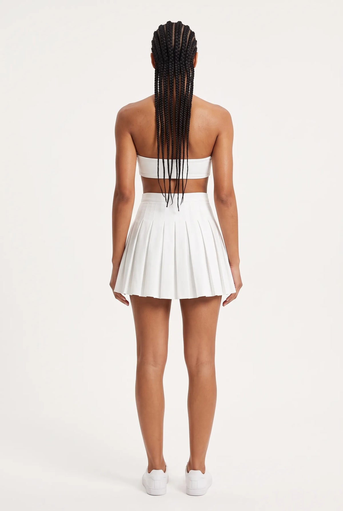 A model viewed from the back wearing a white two-piece outfit consisting of a strapless bandeau-style crop top with a horizontal back cutout and a matching high-waisted pleated mini skirt, paired with white sneakers against a plain off-white studio background.