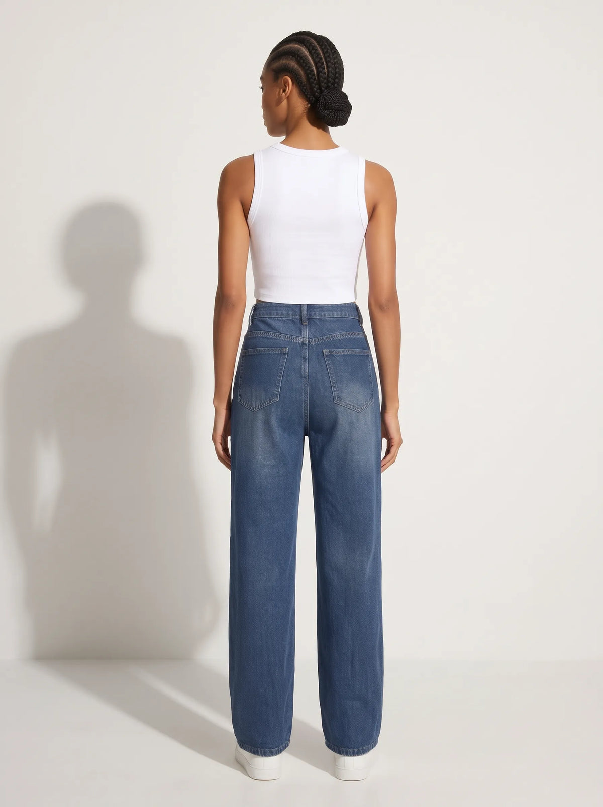 a model wearing a pair of high-waisted, wide-leg denim jeans crafted from a medium-to-dark classic indigo blue wash with a subtle vertical fade down the legs and faint whiskering across the lap. The structured design features a classic five-pocket configuration, belt loops, contrast topstitching throughout, and a secure front closure with a single metallic top button and a central zip-fly.