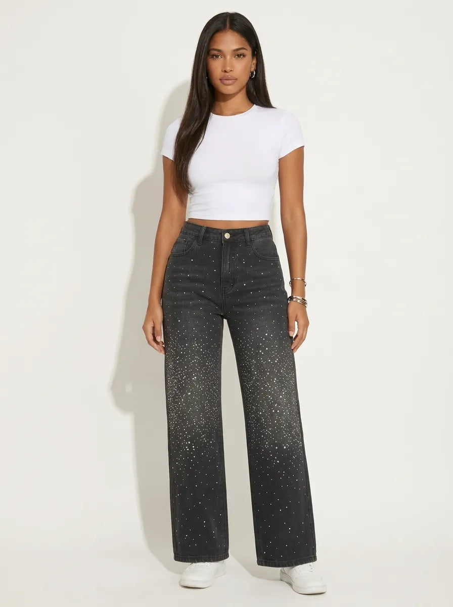 A model wearing a pair of high-waisted, wide-leg jeans is crafted from a dark washed-black or washed-charcoal denim completely covered in a sparkling, all-over pattern of tiny silver rhinestones and crystal studs that scatter densely down the legs. 