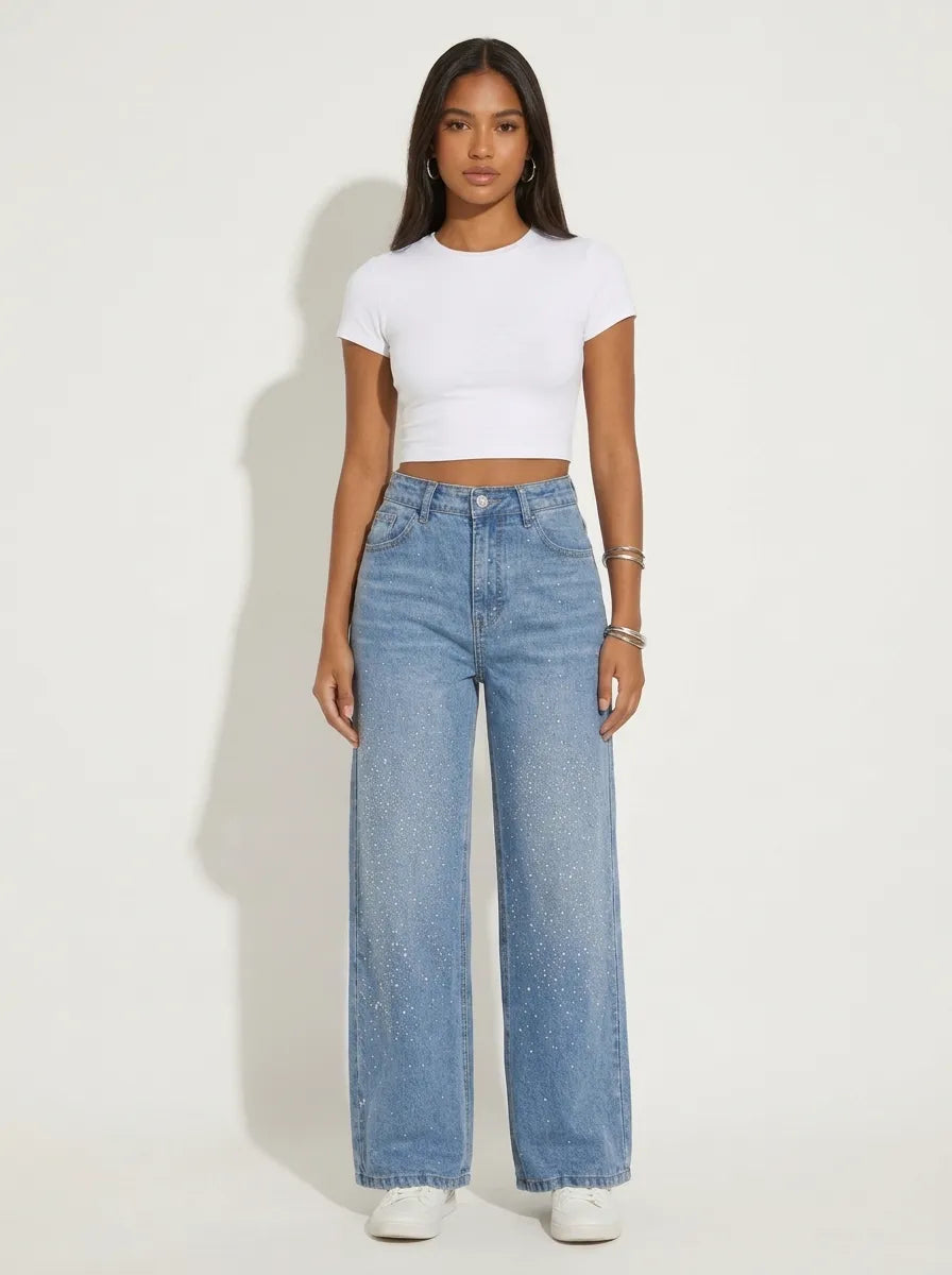 A model wearing a pair of high-waisted, wide-leg jeans is crafted from a washed denim completely covered in a sparkling, all-over pattern of tiny silver rhinestones and crystal studs that scatter densely down the legs. 