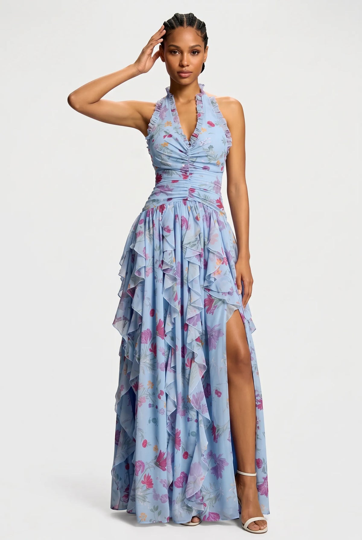 A model wearing a light blue halter-neck maxi dress with a pink and purple floral print, featuring a ruffled tiered skirt and a high side slit, set against a plain light gray background.