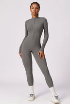 A woman modeling a fitted, long-sleeve gray one-piece jumpsuit with a half-zip neckline, standing against a plain light gray studio background.