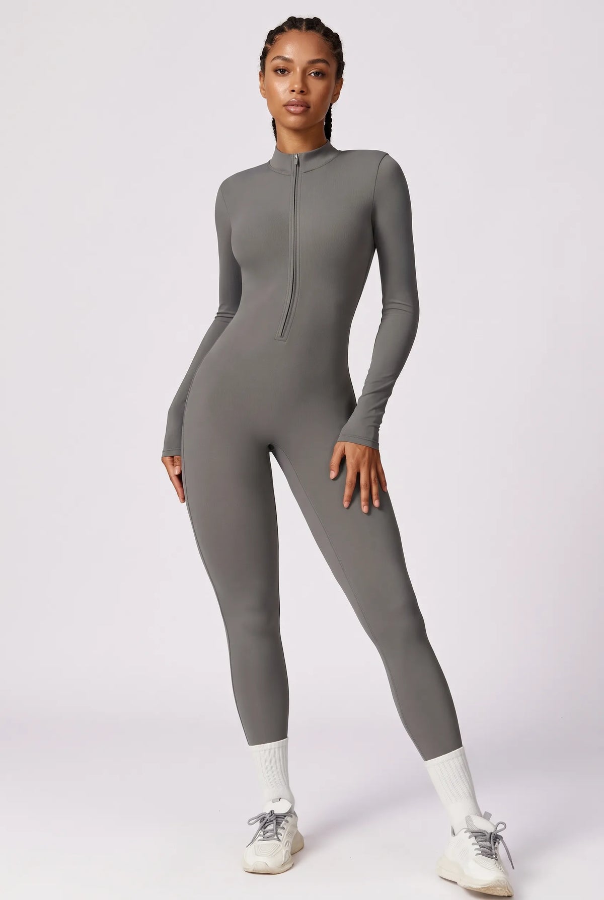 A woman modeling a fitted, long-sleeve gray one-piece jumpsuit with a half-zip neckline, standing against a plain light gray studio background.