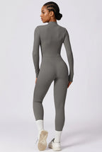 A woman modeling a tight-fitting, long-sleeve gray mock-neck jumpsuit from the back, paired with white socks and white sneakers, against a light gray studio background.