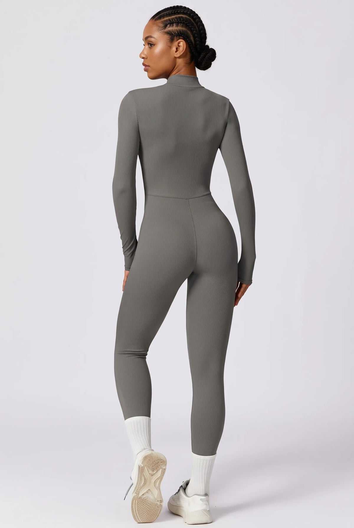 A woman modeling a tight-fitting, long-sleeve gray mock-neck jumpsuit from the back, paired with white socks and white sneakers, against a light gray studio background.