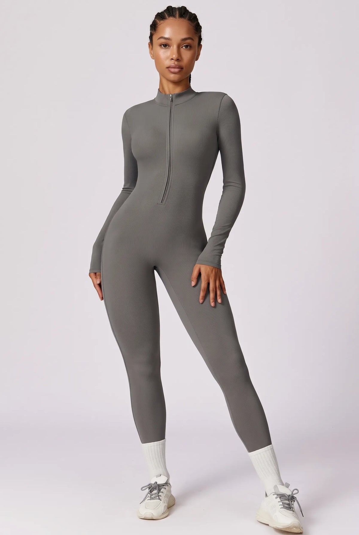 A model wearing a fitted, long-sleeve gray one-piece jumpsuit with a mock neck and front zipper, styled with white socks and white sneakers against a light gray studio background.