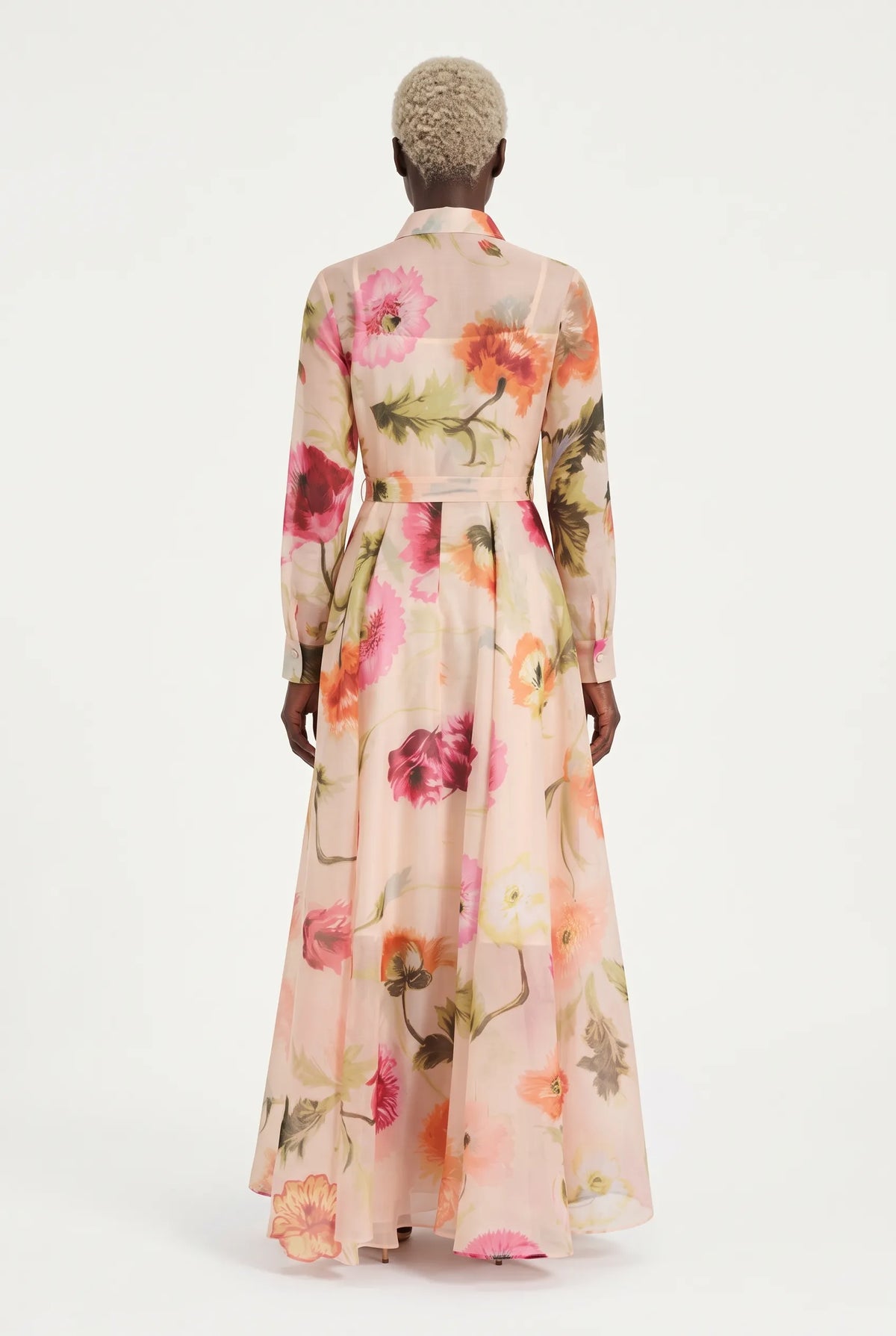 A model wearing a long-sleeved, button-up maxi dress with a pink, peach, and orange floral print on a light beige base, set against a plain white studio background.