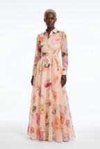 A model wearing a long-sleeved, collared maxi dress with a pink and peach floral print, standing against a plain white studio background.