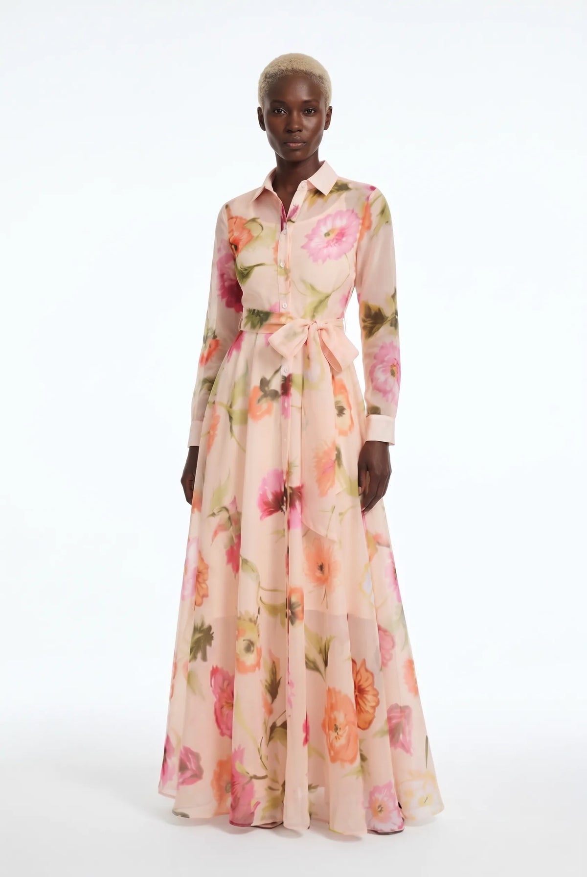 A model wearing a long-sleeved, collared maxi dress with a pink and peach floral print, standing against a plain white studio background.