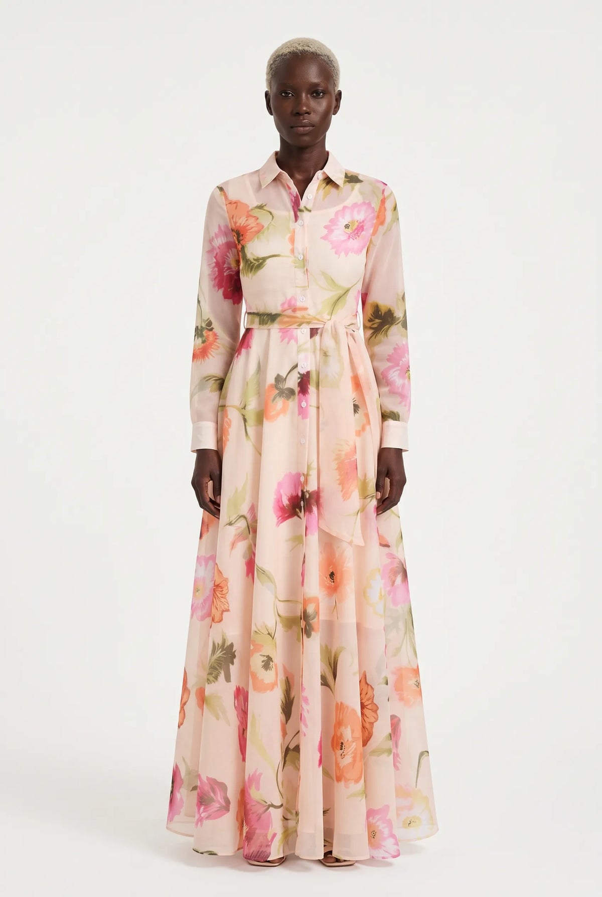 A model wearing a long-sleeved, button-up maxi dress with a pink, peach, and orange floral print on a light beige base, set against a plain white studio background.