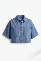 This is a medium-wash denim short-sleeved cropped jacket features a boxy, modern silhouette with relaxed drop shoulders and wide, elbow-length sleeves with stitched turned hems.