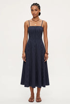 A woman modeling a navy blue midi dress with thin vertical white pinstripes, featuring spaghetti straps and a buttoned bodice, set against a plain light gray studio background.