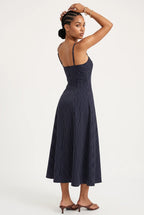 A woman modeling a navy blue midi dress with thin vertical pinstripes, shown from a three-quarters back view against a plain white studio background.