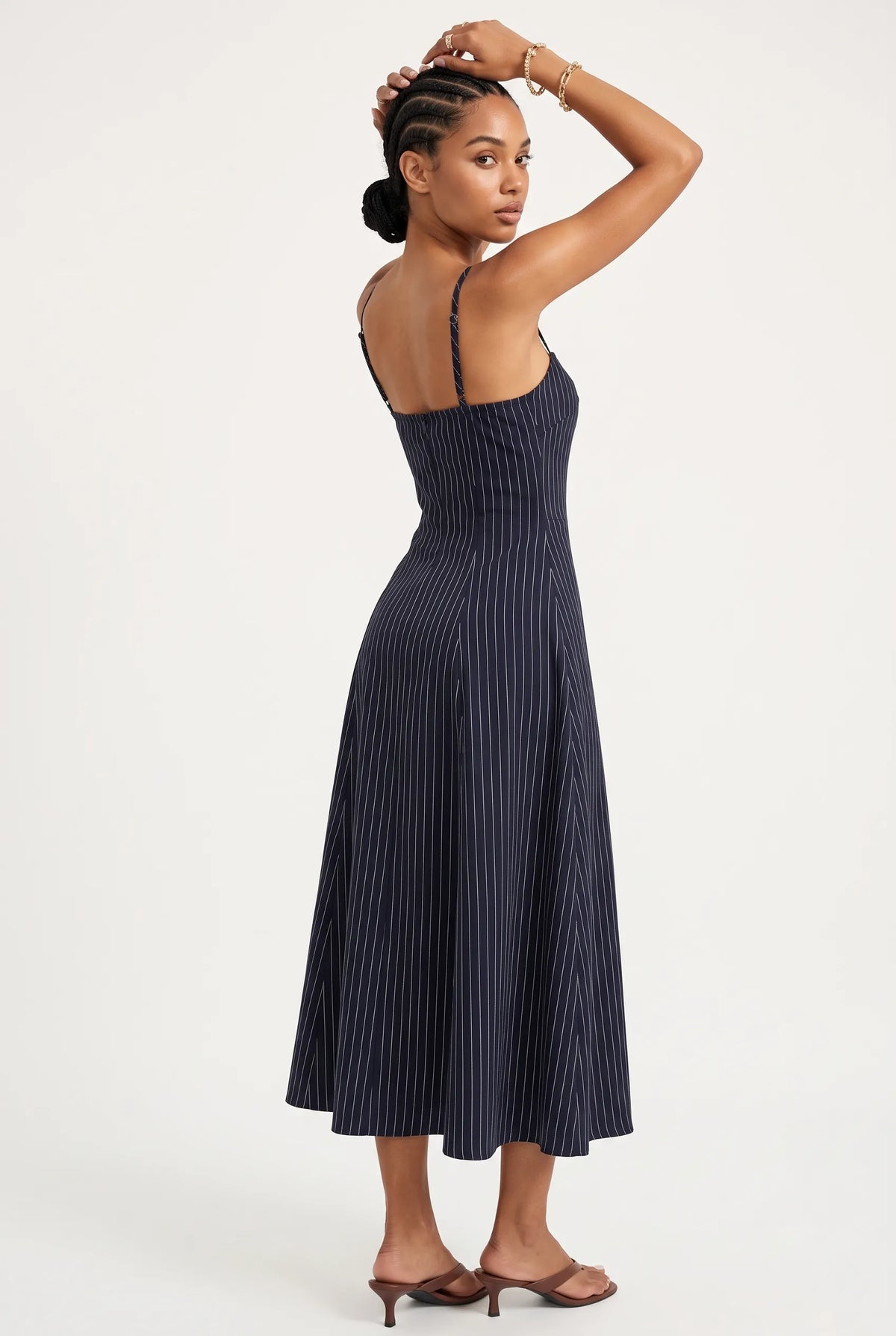 A woman modeling a navy blue midi dress with thin vertical pinstripes, shown from a three-quarters back view against a plain white studio background.
